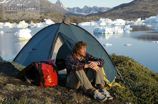 Junger Mann  Abenteurer sitzt vorm Zelt  hinten Eisberge  Paarnakajiit  Sermilik Fjord  Ostgrönland  Grönland  Arktis