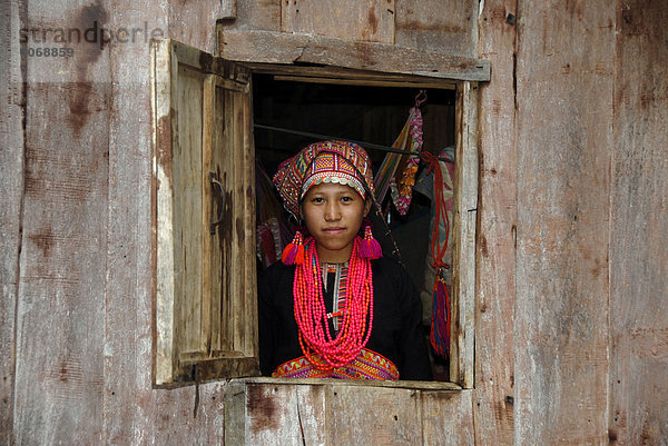 Junge Frau der Akha Pala Ethnie schaut aus dem Fenster bekleidet mit buntem Kopfschmuck und roten Halsketten  Ban Saenkham Tai  Phongsali Provinz  Laos  Südostasien