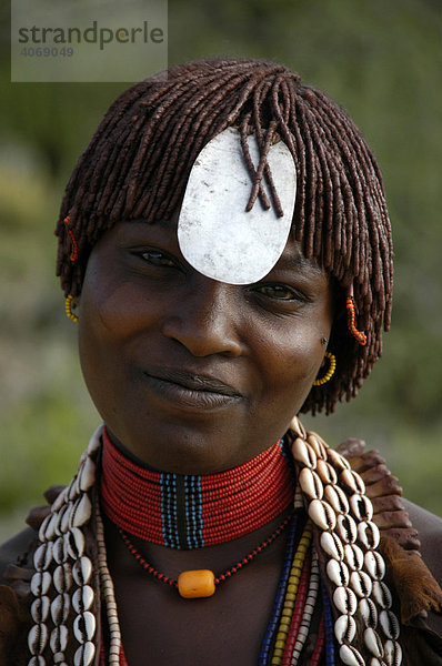 Portrait  junge Frau lächelt mit Kette aus Kaurimuscheln  Metallplättchen vor der Stirn und Lehm in den Haren  bei Turmi  Äthiopien  Afrika