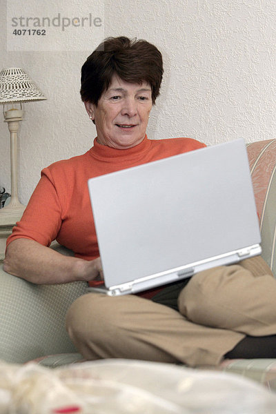 Frau  Seniorin  sitzt im Wohnzimmer auf einem Sofa vor einem Laptop