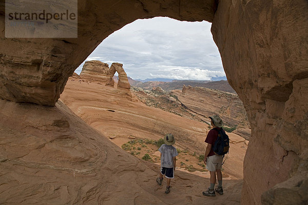 John West und sein Sohn Joey  9  unter einem kleinen Felsenbogen mit Blick auf den Delicate Arch Felsbogen  Arches Nationalpark  Moab  Utah  USA