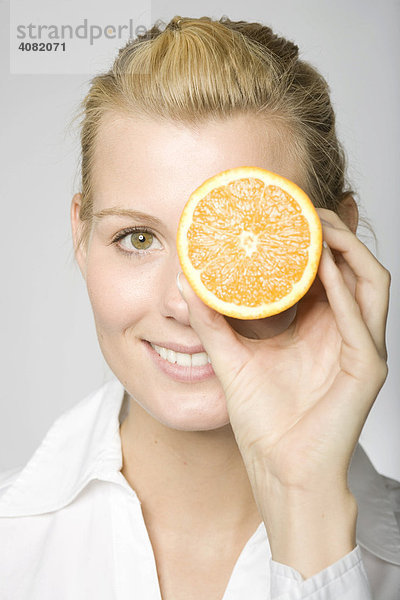 Blonde Frau hält sich eine aufgeschnittene Orange vor ein Auge und lacht
