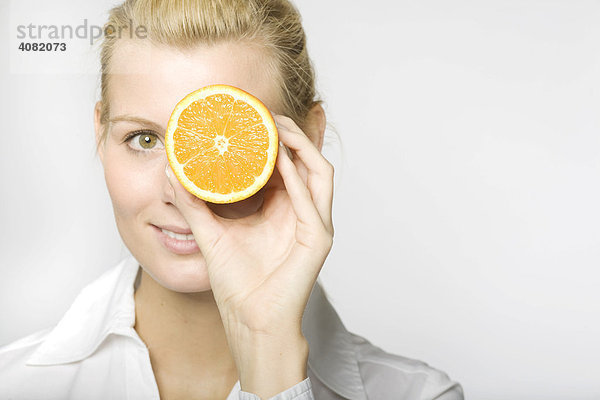 Blonde Frau hält sich eine aufgeschnittene Orange vor ein Auge und lacht