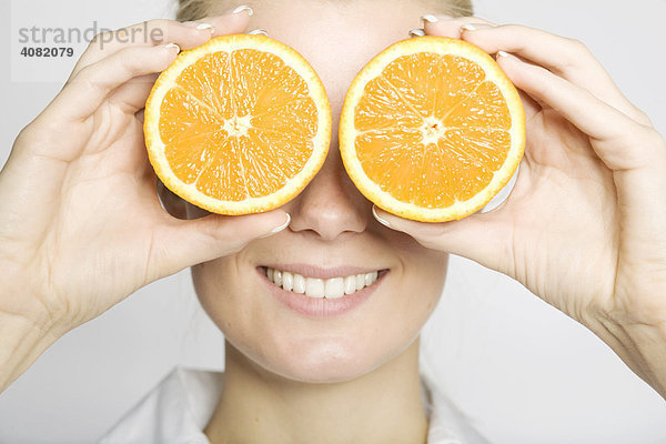 Blonde Frau hält sich zwei aufgeschnittene Orangen vor die Augen und lacht
