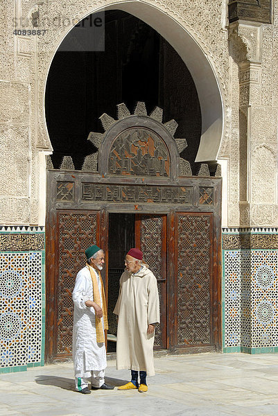 Männer in traditioneller Kleidung stehen vor einem reich verzierten Tor aus Holz und reden miteinander Medersa Bou Inania Fes El-Bali Marokko