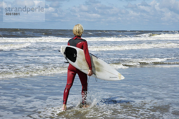 Blonder junger Mann in einem rotem Neoprenanzug trägt Surfbrett am Strand  Katwijk aan Zee  Südholland  Holland  Niederlande