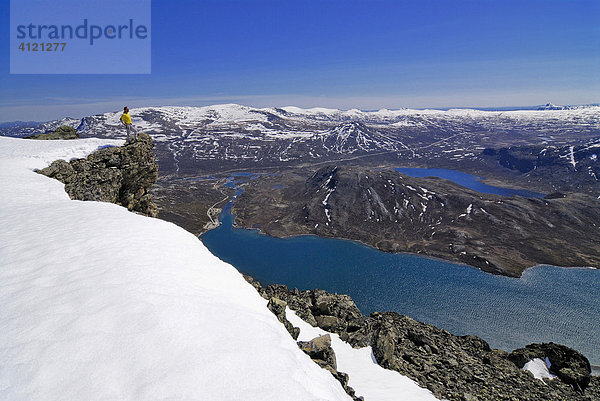 Wanderer blickt von einer verschneiten Felskanzel über eine weite Landschaft mit Bergen und Seen  Jotunheimen Nationalpark  Vaga  Oppland  Norwegen