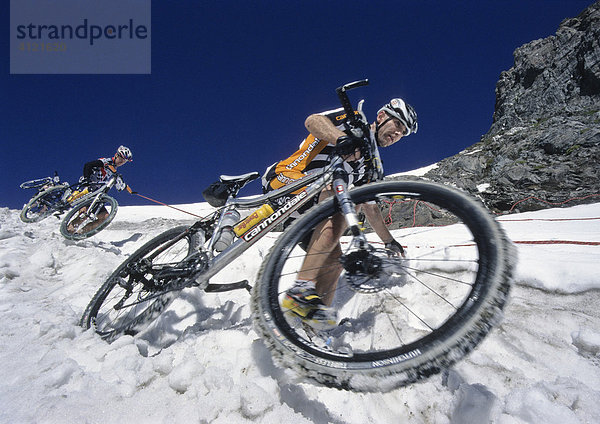 Zwei Teilnehmer der Adidas Bike Transalp Challenge  Mountainbikerennen  überqueren einen verschneiten Bergpass und tragen ihre Bikes über den Schnee hinunter  Pfunderer Joch  Südtirol  Italien  Europa