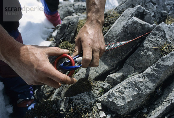 Männerhände haken einen Abseilachter in einen Alukarabiner der an einer Bandschlaufe am Fels befestigt ist