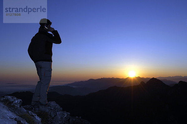 Mann mit Handy steht bei Sonnenaufgang auf einem Berggipfel  Breitenstein  Bayerische Voralpen  Wendelsteingruppe  Oberbayern  Bayern  Deutschland