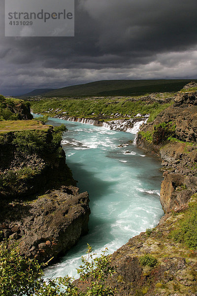 Hvita-Fluss mit seitlich rechts eintretenden Lavawasserfällen (Hraunfossar-Wasserfälle) nahe beim Langjökull-Gletscherberg mit dunklen Gewitterwolken in Island