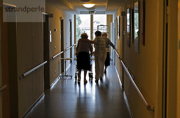 Eine Pflegerin hilft alter Frau mit Gehhilfe (Rollator) über den Flur.