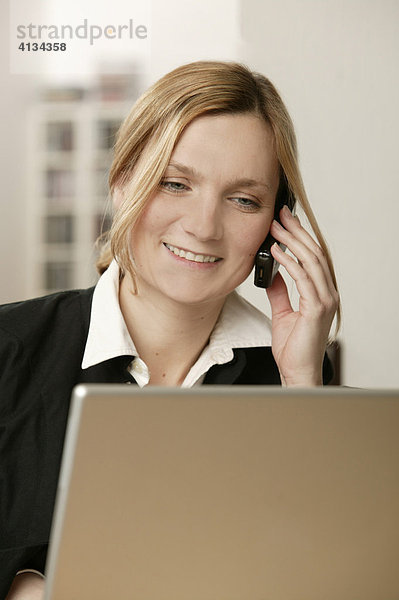 Junge blonde Frau am Laptop mit Telefon  lächelt