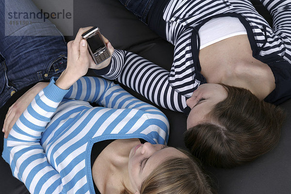 Zwei Mädchen liegen auf großem Kissen und versenden SMS