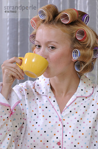 Rothaarige Frau mit Lockenwicklern im Haar und Pyjama trinkt aus einer gelben Tasse