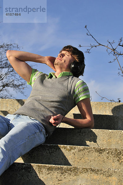Junger Mann sitzt auf einer Treppe unter freiem  blauem Himmel und hört mit Kopfhörern Musik