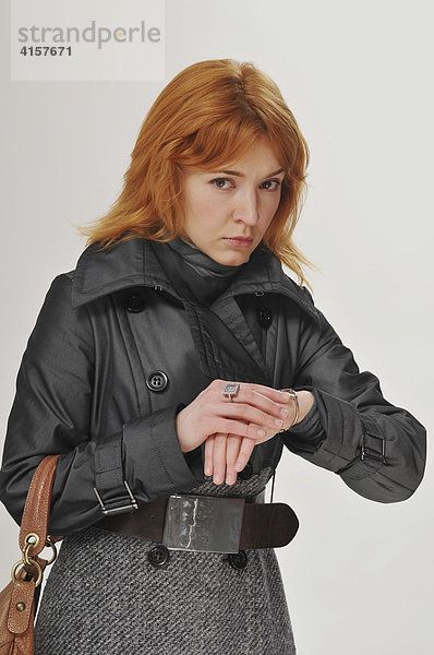 Rothaarige Frau mit grauem Mantel und rotbrauner Tasche schaut auf die Uhr