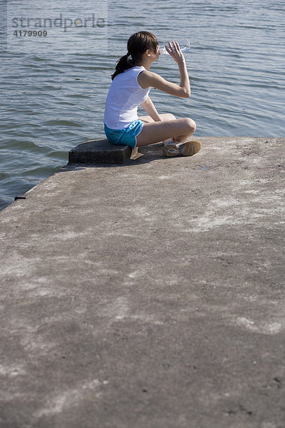 Junge Sportlerin sitzt am Wasser und trinkt aus einer Flasche