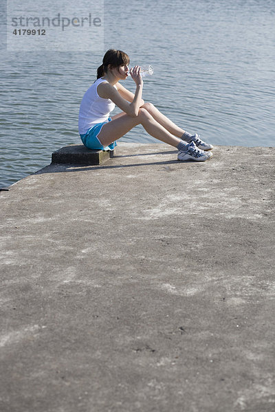 Junge Sportlerin sitzt am Wasser und trinkt aus einer Getränkeflasche