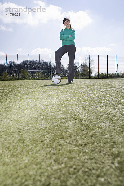 Junge Frau steht auf einem Sportplatz und hat ein Bein auf den Ball gestellt