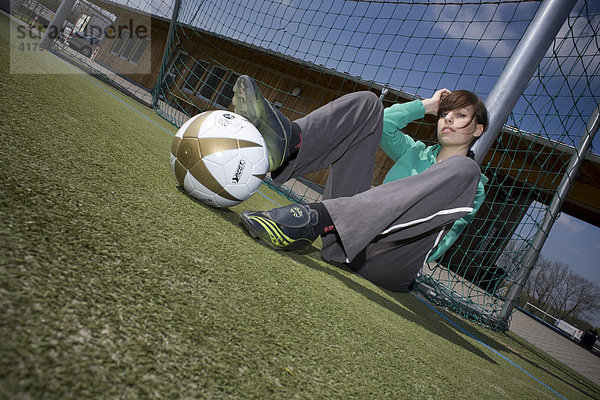 Junge Frau sitzt mit einem Fußball an einem Torpfosten gelehnt