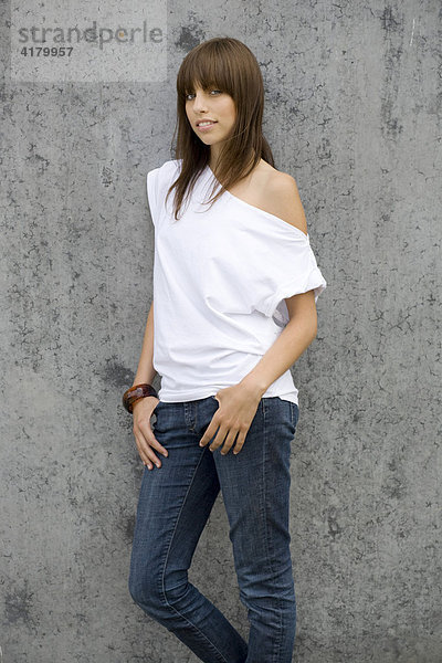 Junge dunkelhaarige Frau steht mit Jeans und weißem Top vor einer Wand und blickt entspannt in die Kamera