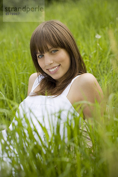 Junge dunkelhaarige Frau in einem weißen Kleid liegt entspannt auf einer Wiese  genießt den Sommer  blickt lächelnd in die Kamera