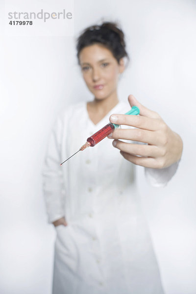 Junge Frau mit weißem Laborkittel hält eine Spritze mit roter Flüssigkeit in die Kamera