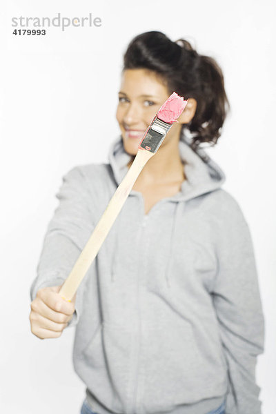 Junge Frau hält einen Pinsel mit pinker Farbe in den Händen und lacht in die Kamera