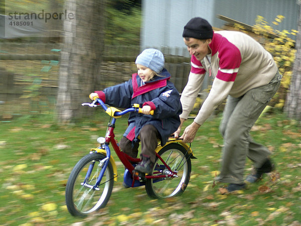 Erste Fahrversuche der vierjaehrigen Mädchen auf dem neuen Fahrrad ohne Stuetzraeder mit Hilfe des Vaters im Garten.