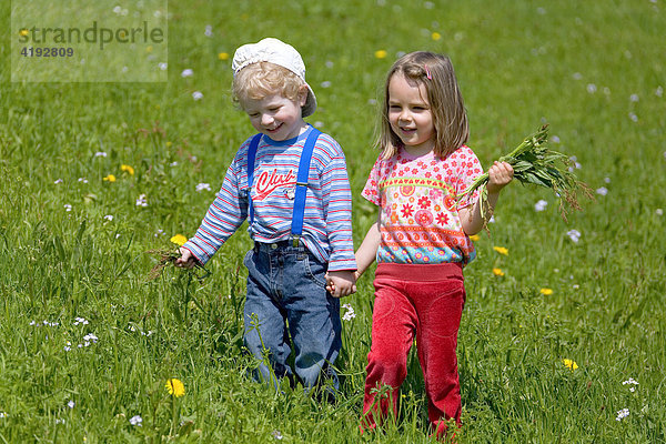 Ein Mädel und eine Bube spazieren Hand in Hand mit Blumen über eine Wiese