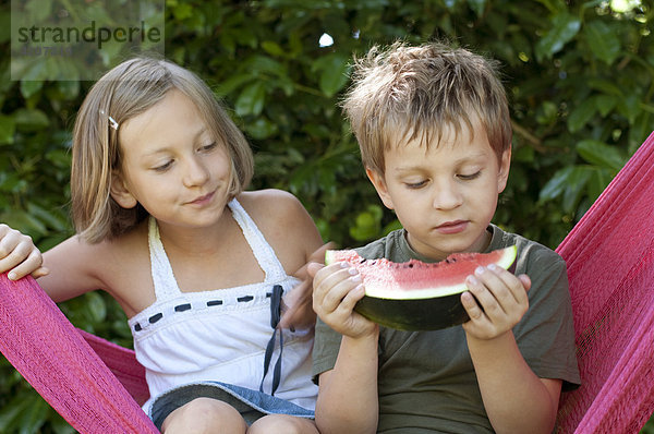 Junge und Mädchen sitzen in einer Hängematte  Junge isst Melone