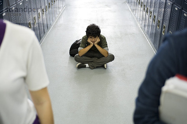 Teenager-Junge sitzt mürrisch auf dem Boden des Schulflurs  Klassenkameraden im Vordergrund