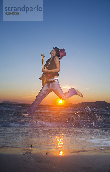 Junger Mann springt mit Gitarre bei Sonnenuntergang