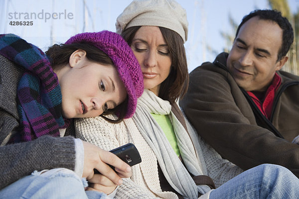 Teenager-Mädchen sitzt mit Eltern im Freien  SMS mit ihrem Handy