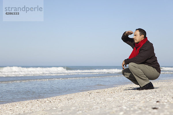 Mann hockt am Strand und schaut auf den Ozean.