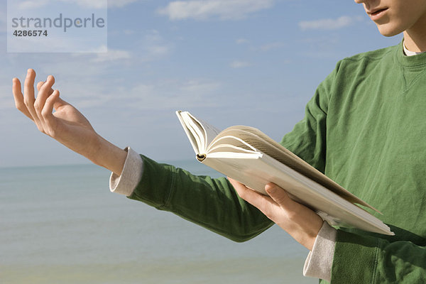 Junger Mann liest laut aus einem Buch am Strand vor