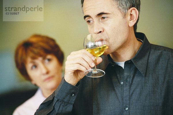 Senior Mann trinkt ein Glas Wein  Seniorin schaut ihn im Hintergrund an.