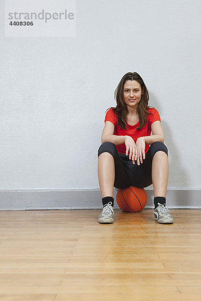 Junge Frau sitzt auf Basketball in der Turnhalle und lächelt.
