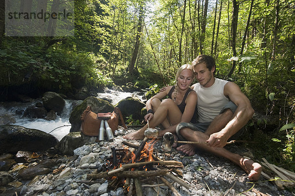 Österreich  Steiermark  Junges Paar sitzt am Lagerfeuer am Bach im Wald