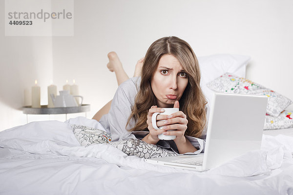 Frau liegt auf dem Bett und hält Kaffeetasse mit Laptop  Stirnrunzeln