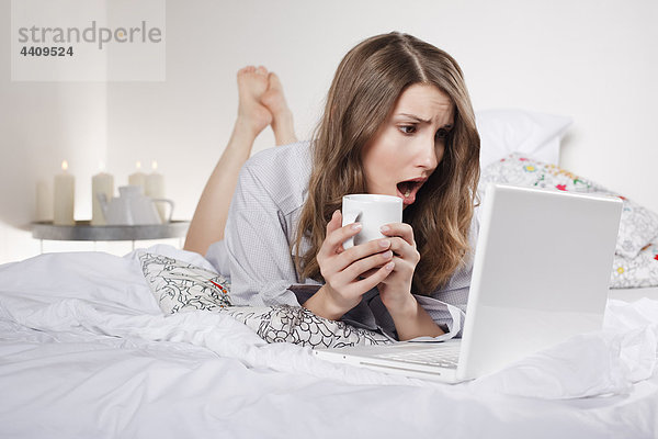 Frau auf dem Bett liegend  hält Kaffeetasse mit Laptop  Mund offen