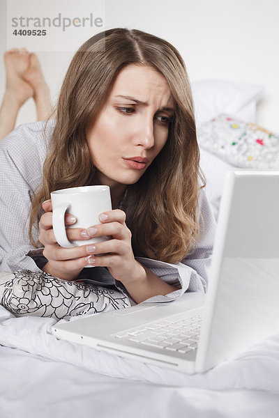 Frau auf dem Bett liegend mit Kaffeetasse und Laptop