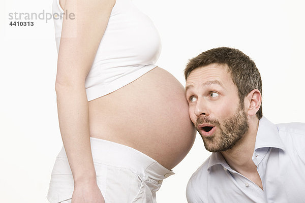Mann  der dem Bauch einer schwangeren Frau lächelnd zuhört.
