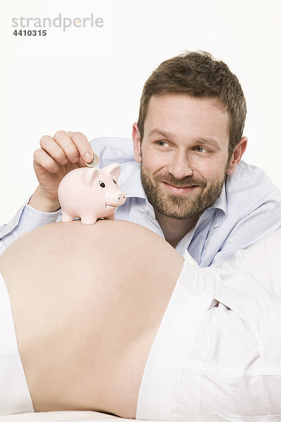 Mann  der Geld in Sparschwein auf den Bauch einer schwangeren Frau legt  Nahaufnahme