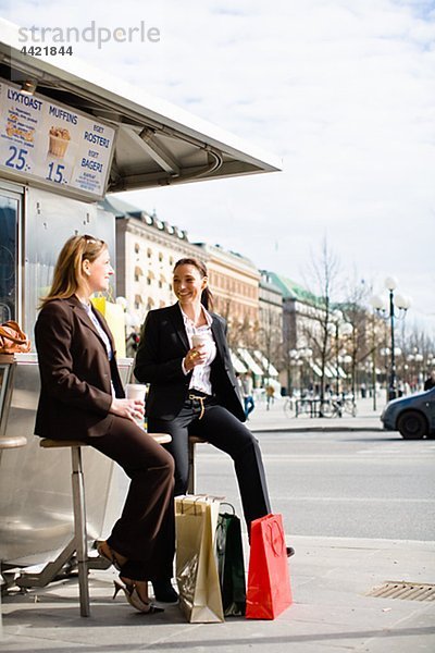 Paar von eleganten Frauen warten auf Bushaltestelle mit Einkaufstaschen