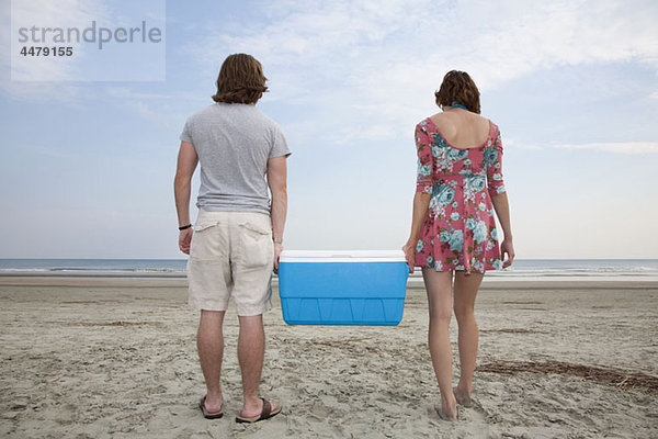 Ein junges Paar trägt eine Kühlbox zusammen am Strand.