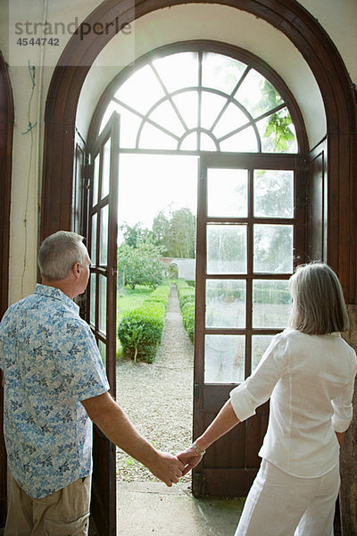 Ein reifes Ehepaar hält sich an einer Fenstertür an den Händen.