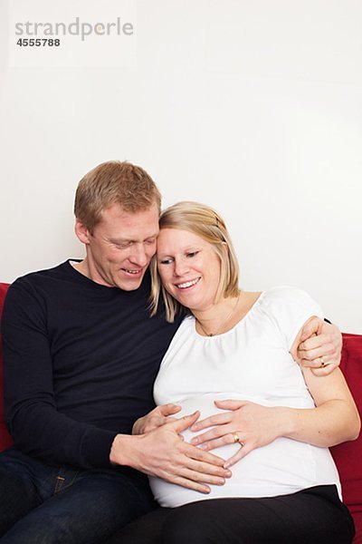Mann mit schwangere Frau sitzt auf Sofa an Hand auf den Bauch