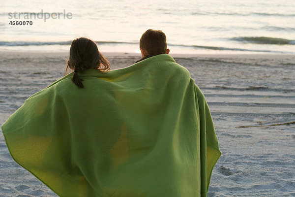 Kinder zusammen in eine Decke gewickelt  die am Strand steht und den Sonnenuntergang beobachtet.